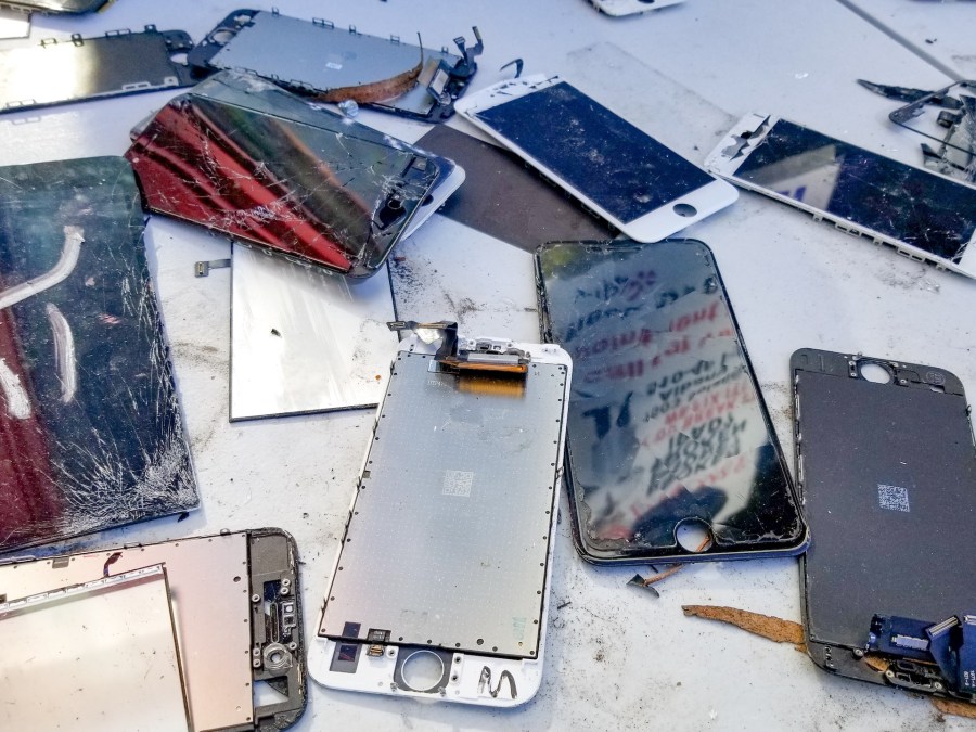 Close-up of a large number of broken cellphones, including cracked cellphone screens with exposed wiring.