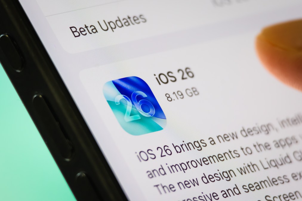 A smartphone screen shows the Apple iOS 26 update with a user's finger preparing to install it. (Photo illustration by Cheng Xin/Getty Images)